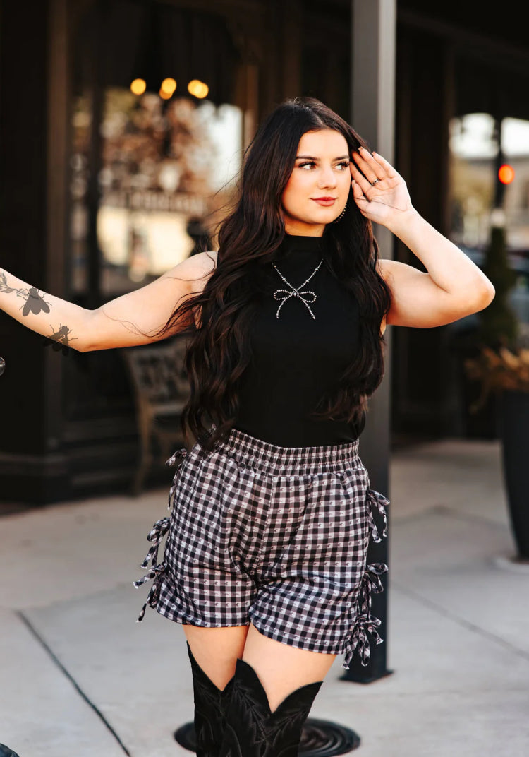 Woman wearing a black top and plaid shorts with cowboy boots, standing on a street.