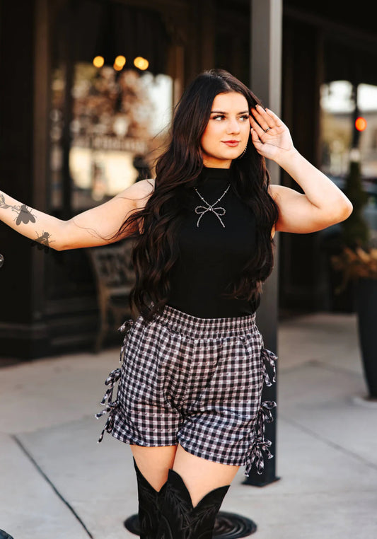 Woman wearing a black top and plaid shorts with cowboy boots, standing on a street.